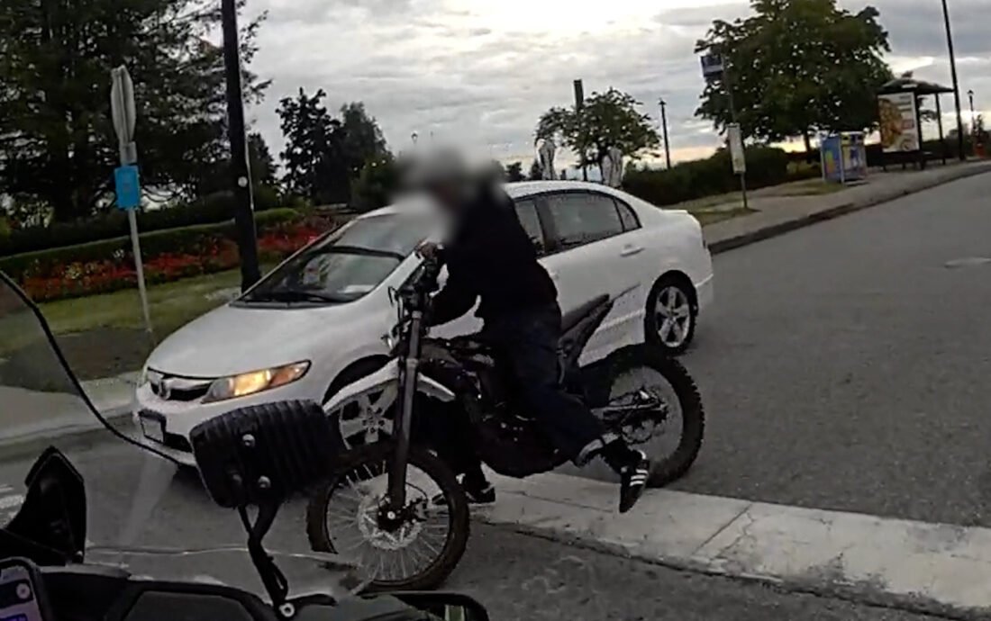 A person rides an electric motorcycle past a parked car, face blurred, in a still from BC Highway Patrol video in North Vancouver