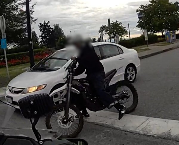 A person rides an electric motorcycle past a parked car, face blurred, in a still from BC Highway Patrol video in North Vancouver